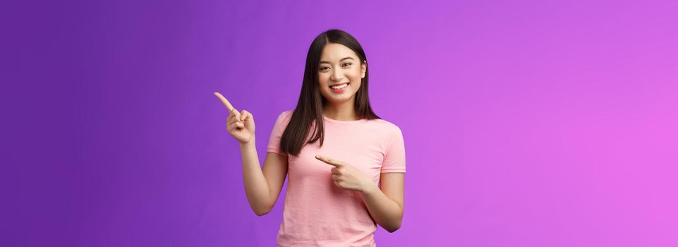 Cheerful friendly tender asian smiling girl pointing right, showing promo, introduce new product, grinning joyfully present cool adverstiment, stand pink t-shirt purple background amused outgoing.