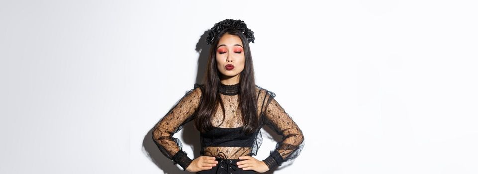 Image of beautiful asian girl in halloween costume close eyes and pouting, waiting for kiss, standing over white background wearing gothic black dress with wreath made of roses.