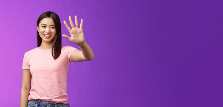 Friendly cute asian tender girl raise palm waving goodbye, smiling joyfully, farewell, gladly welcome friends inviting come in, saying hi hello, greeting, stand purple background. Copy space