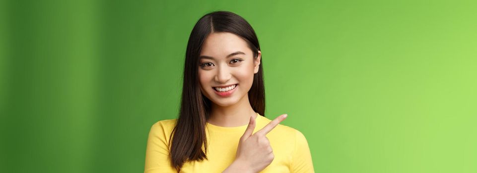 Cheerful good-looking asian brunette showing excellent promo opportunity, pointing index finger left, smiling glad, pleasantly help introduce product, stand green background. Copy space
