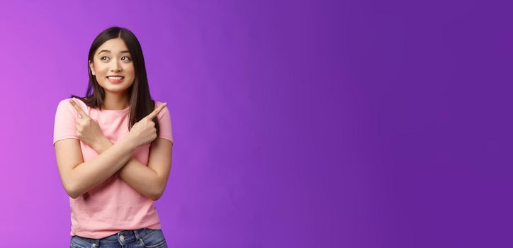 Cheerful timid cute asian girl, look tender, smiling gazing aside cross hands chest, pointing sideways, show left right, making decision, gathering confidence choose, purple background.