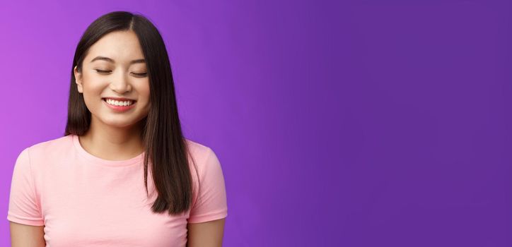 Cheerful tender lovely asian girl brunette, close eyes happily smiling, satisfied, receive good news, dream come true, stand purple background joyful, grinning from happiness and joy.