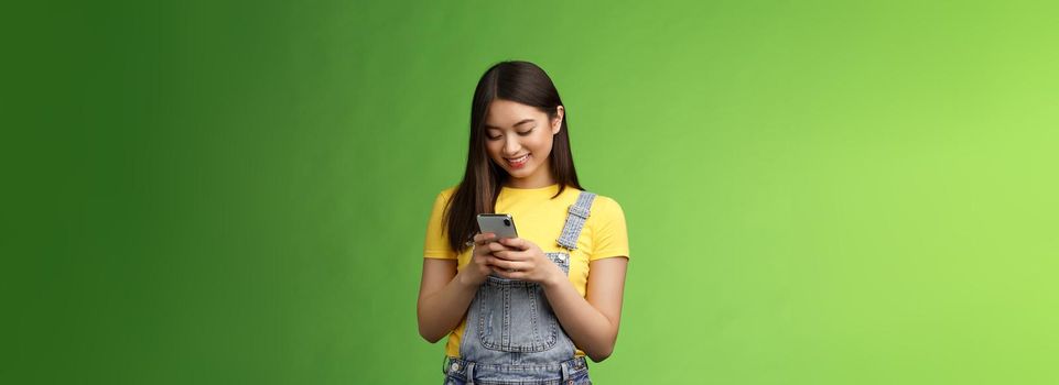 Outgoing cute asian brunette woman scroll online shop, purchase summer tour internet, hold smartphone, look amused telephone screen, use app, edit photos post social media, stand green background.