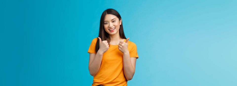 Supportive cute friendly asian girl give thumbs up and smiling, cheering for you, congratulate nice work, approving good idea, like nice outfit, grinning glad accept plan, stand blue background.