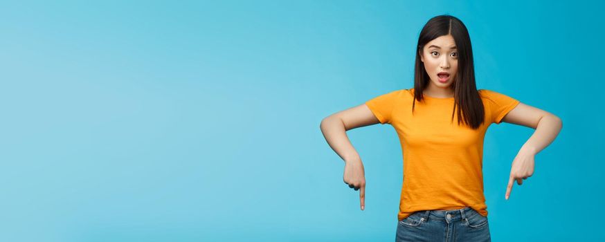 Impressed asian girl drop jaw shocked look camera surprised pointing down indicaring bottom advertisement, gasping open mouth astonished curious about interesting opportunity, blue background.