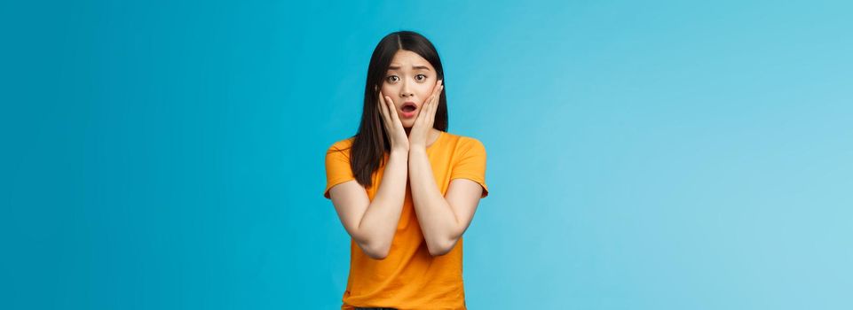 Shocked concerned young asian girl look empathy camera, gasping open mouth worried, frowning upset hear frustrating news, pitty feel sorry for friend, touch cheeks speechless, stand blue background.