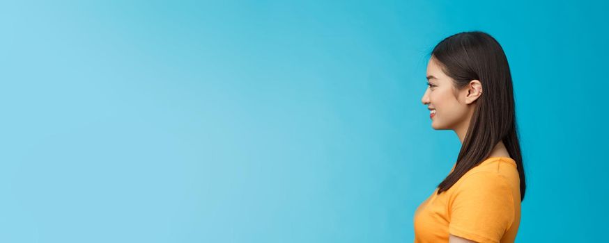 Profile shot asian cute woman perfect skin, turn left, smiling joyfully, pose near blue background, standing queue, awaiting coffee take-away, relaxed chatting girlfriend, wear yellow summer t-shirt.