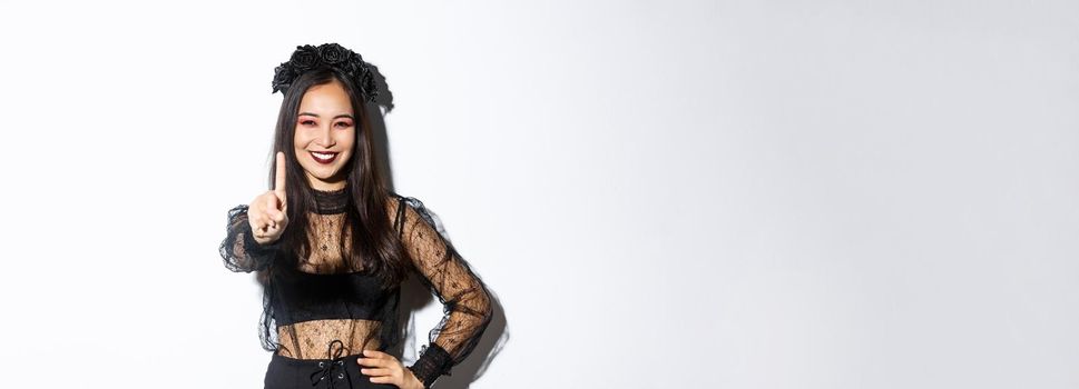 Image of smiling beautiful asian woman in halloween costume showing stop gesture, extend one finger and looking happy, prohibit or reject something, standing white background.