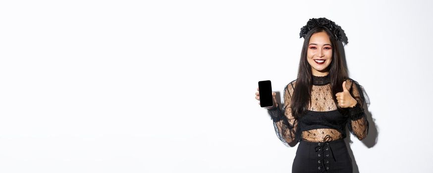 Image of happy and satisfied asian woman in halloween costume showing thumbs-up and demonstrating mobile phone screen, smiling pleased, standing over white background.