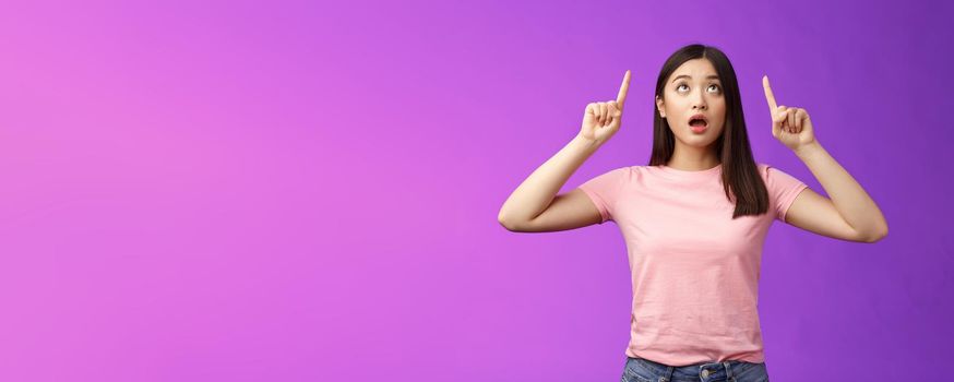 Surprised impressed asian girl brunette look fascinated up, drop jaw gasping astonished, check out impressive interesting promo, pointing top advertisement amazed, stand purple background.