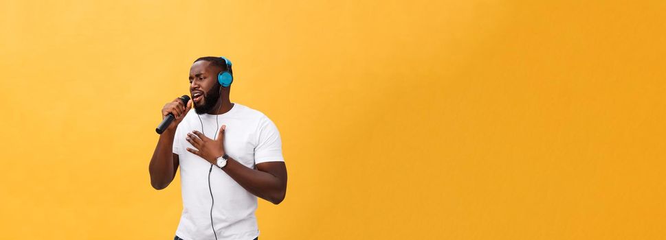 Portrait of cheerful positive chic. handsome african man holding microphone and having headphones on head listening music singing song enjoying weekend vacation isolated on yellow background.