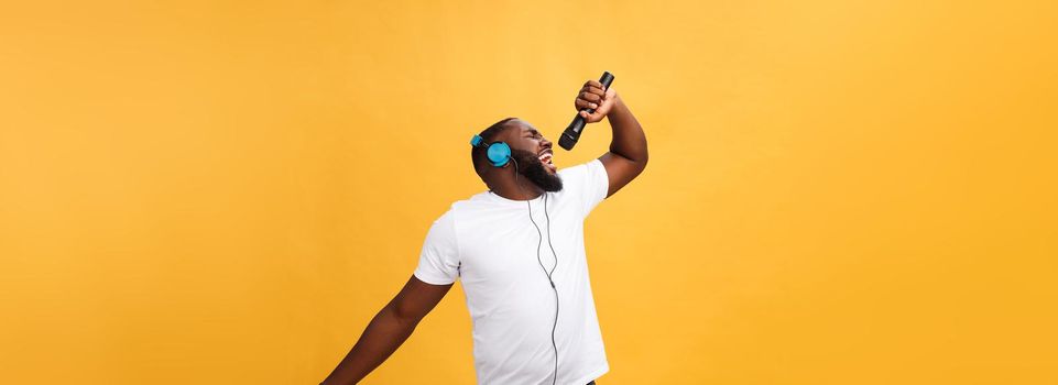 Portrait of cheerful positive chic. handsome african man holding microphone and having headphones on head listening music singing song enjoying weekend vacation isolated on yellow background.