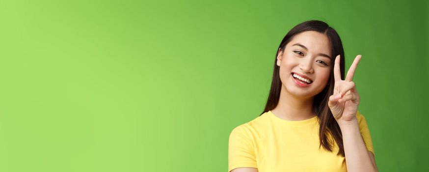 Close-up joyful cute asian girl lively look camera smile, show peace victory sign entertained, grinning posing photograph tender expression, stand green background positive and lucky.