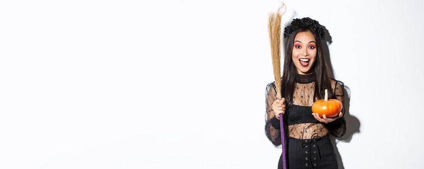 Excited attractive asian girl celebrating halloween, wearing witch costume, holding pumpkin and broom, going trick or treat, standing over white background.