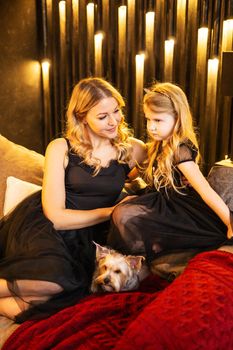 Mom and daughter in festive attire on the couch with their dog next to a Christmas tree decorated with garlands, balloons and Christmas toys. The concept of winter holidays is Christmas and New Year holidays. Magical festive atmosphere.