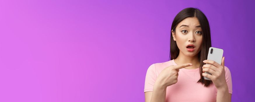 Concerned confused cute young asian woman hold smartphone open mouth, pointing finger telephone, look questioned upset, troubled see strange post online, stand purple background. Copy space