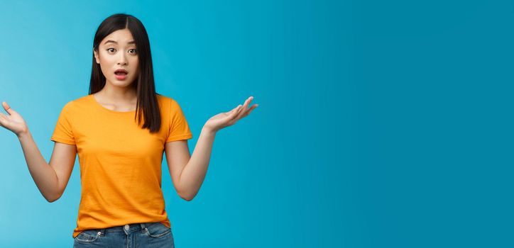 Questioned unaware young asian girl with dark short haircut shrugging hands spread sideways, look confused open mouth uncertain, being clueless, stand blue background wear yellow t-shirt.