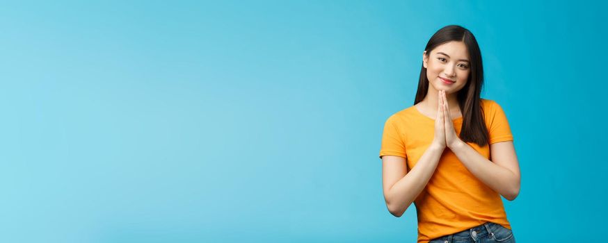 Pretty please girl asking politely. Cute charming asian woman smiling tenderly, hold hands pray grinning begging for favour, plead, look thankful for help stand blue background joyful, caring.