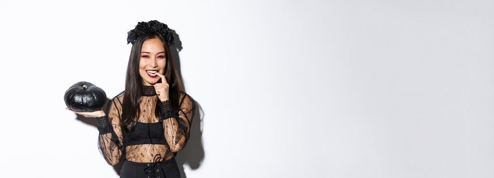 Sassy attractive asian woman in witch costume looking cunning and smiling, holding pumpking, going trick or treating on halloween, standing over white background.