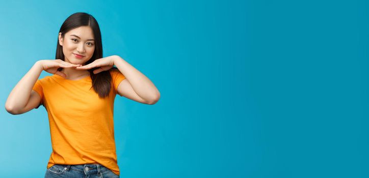 Sweet lovely tender asian girl brunette, tilt head smiling show clean pure face hold palms under jawline satisfied after applying skincare product stand blue background wear yellow t-shirt.