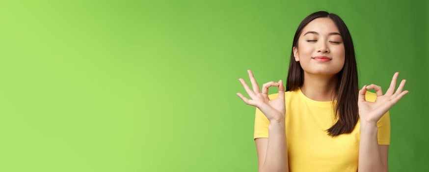 Peaceful charming asian girl brunette inhale fresh air, relaxing breathing happily, close eyes smiling, hold hands zen mudra signs reach nirvana, meditation, practice yoga, green background.