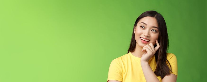 Close-up dreamy intrigued creative asian brunette woman imaging summer vacation trip, picturing perfect holiday, standing thoughtful, touch cheek, thinking look up, stand green background.