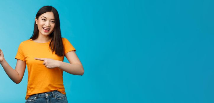 Upbeat friendly asian cute girl with dark haircut lively pointing left introduce awesome new copy space, smiling broadly toothy happy grin, stand blue background joyful, recommend link.