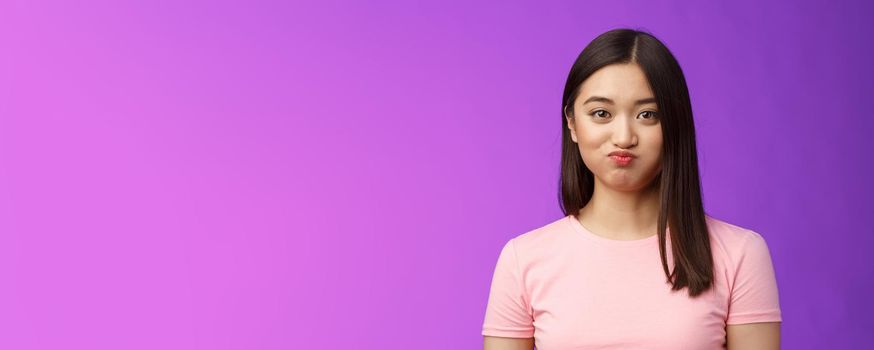 Close-up cute tender young asian female holding breath speechless, smiling silly having challenge hiding secret unwilling talk, pouting lovely, look camera entertained, purple background.