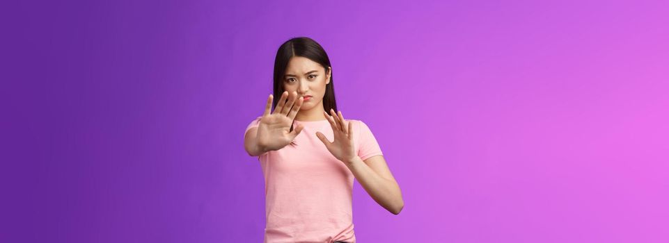 Displeased intense reluctant asian girl blocking, trying protect face, raise hands stop, prohibition gesture, frowning sulking, refusing bothering offer, stand purple background, rejecting.