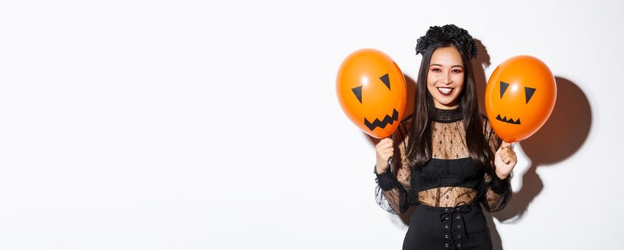 Image of cheerful asian woman in witch costume celebrating halloween, holding balloons with scary faces, standing over white background.