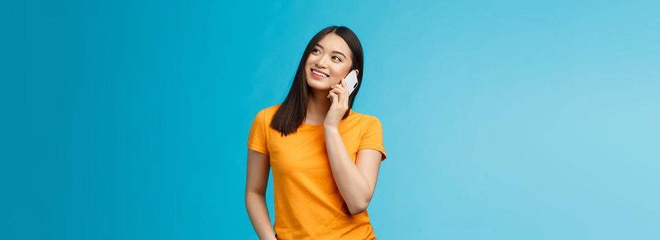 Friendly cute urban asian girl look up dreamy relaxed, smiling joyfully hold smartphone near ear, talking casually friend via telephone, satisfied good mobile connection, stand blue background.