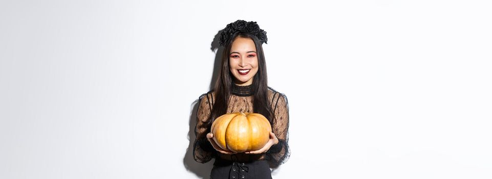 Happy beautiful woman in black lace dress enjoying halloween holiday, smiling at camera and holding pumpkin, standing over white background.