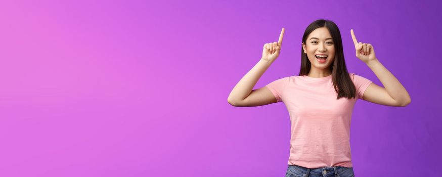 Charismatic good-looking lively smiling asian girl introduce product, pointing fingers up, indicate top advertisement, grinning toothy, excited telling good news, sharing link, purple background.