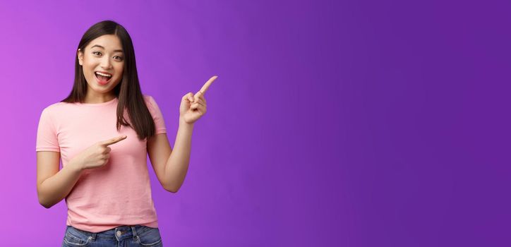 Friendly enthsuasitic amazed attractive tender asian girl having interesting conversation, discuss interest new store, pointing right inviting come inside, stand purple background smiling camera.