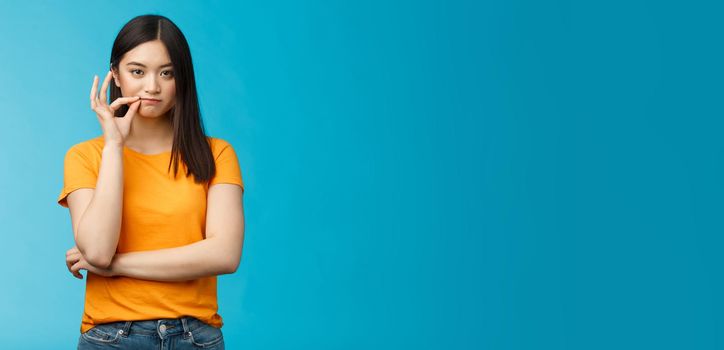 Serious-looking asian female friend promise keep secret, seal lips, hold zip near mouth look focused determined, stay silent and speechless, stand wearing yellow t-shirt blue background.