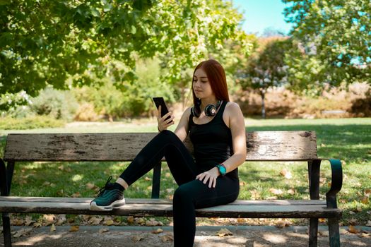 young redhead girl, looking at the smartphone, with music headphones sitting on a park bench