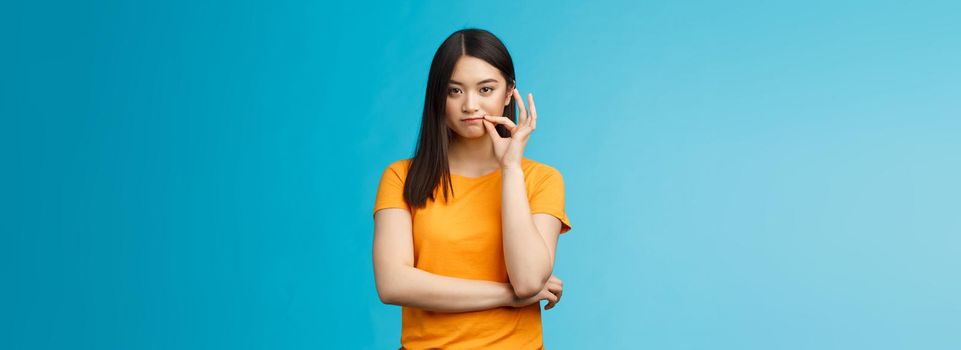 Serious-looking asian female friend promise keep secret, seal lips, hold zip near mouth look focused determined, stay silent and speechless, stand wearing yellow t-shirt blue background.