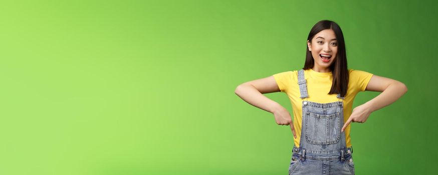 Lively energized asian cute female promote advertisement pointing fingers down, show you cool place, smiling camera joyful, telling great pleasant news stand green background emotional.