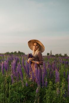 A beautiful woman in a straw hat walks in a field with purple flowers. A walk in nature in the lupin field.