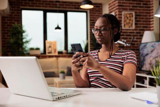 Young freelancer typing message on smartphone, chatting in social network at workplace. Attractive remote worker having break, surfing internet on mobile phone, sitting at desk