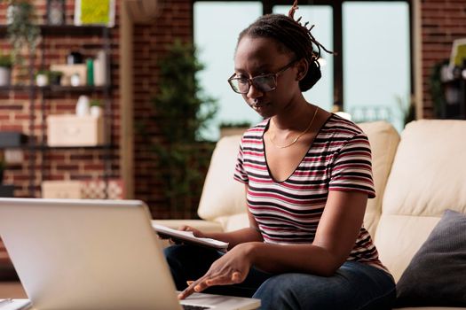 Remote employee holding clipboard with report, working in home office, using laptop. Serious professional freelancer looking at computer, typing, busy remote employee in glasses