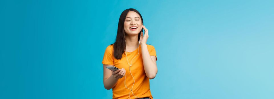 Lovely carefree cute asian girl enjoy awesome music boost mood listening favorite songs, touch earphone close eyes from pleasure delight, singing along, hold smartphone stand blue background.