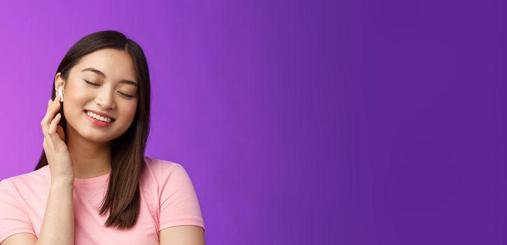 Cute asian girl enjoying listening music, put on wireless headphone, tilt head close eyes dreamy smiling satisfaction, perfect sound, make volume earphone louder, stand purple background.