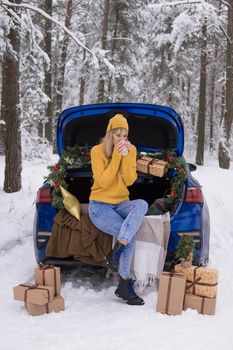 A woman in a winter snow-covered forest in the trunk of a blue car decorated with Christmas decor. The concept of Christmas and winter holidays.