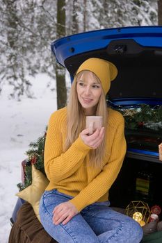 A woman in a winter snow-covered forest in the trunk of a blue car decorated with Christmas decor. The concept of Christmas and winter holidays.