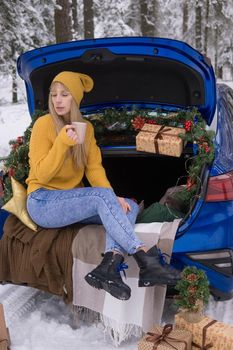 A woman in a winter snow-covered forest in the trunk of a blue car decorated with Christmas decor. The concept of Christmas and winter holidays.