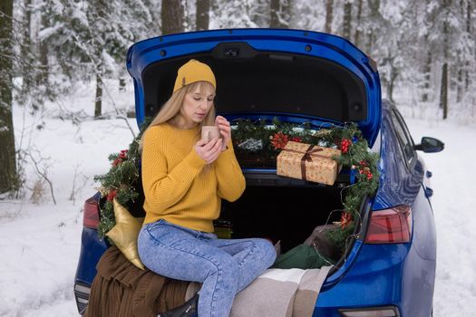 A woman in a winter snow-covered forest in the trunk of a blue car decorated with Christmas decor. The concept of Christmas and winter holidays.