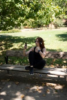 young redhead girl, looking at the smartphone, with music headphones sitting on a park bench