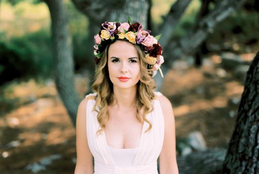 Bride in a wreath of multi-colored flowers on her head stands near a tree. High quality photo