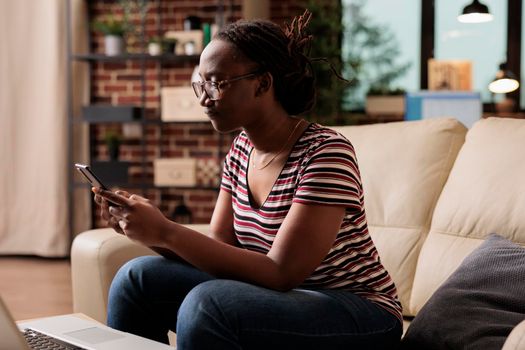 Young african american woman using smartphone, typing message, browsing social media. Freelancer having break, surfing internet on mobile phone, sitting on couch in home room